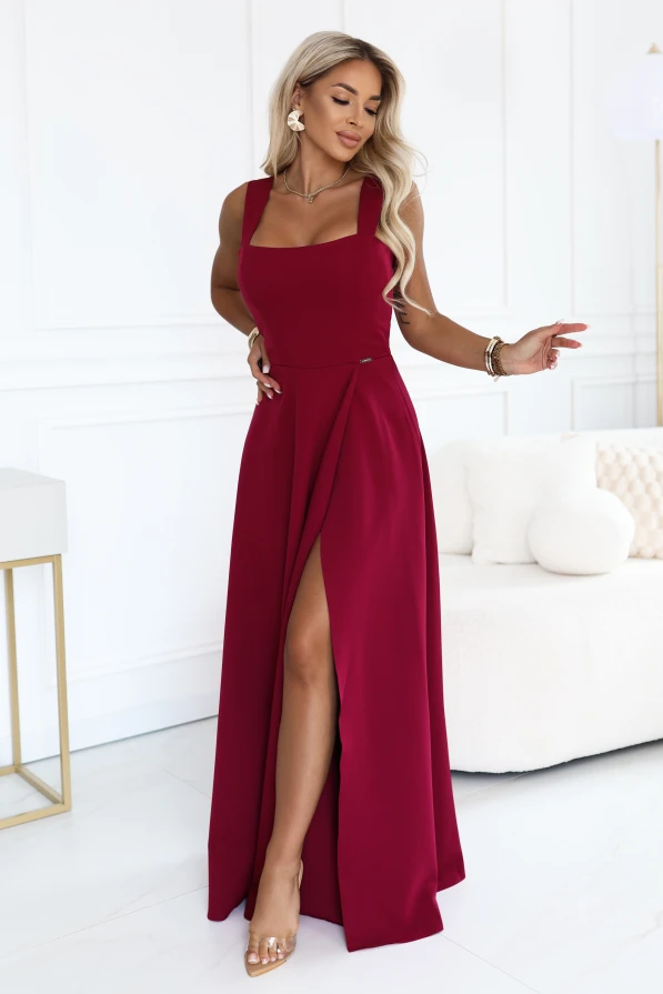 Modelis su elegantiška bordo maxi suknele Numoco, kvadratine iškirpte, plačiomis petnešėlėmis, pabrėžtu juosmeniu ir aukšta įpjova ties koja, pristatoma šviesioje, modernioje erdvėje.