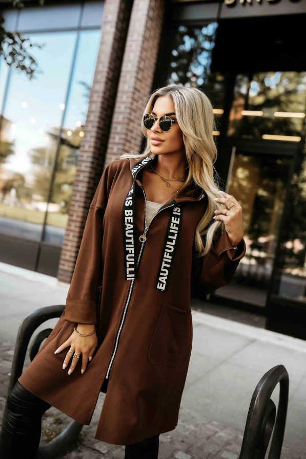 Modelka prezentuje czekoladowy płaszczyk damski z kapturem i kieszeniami marki Numoco Basic, idealny na jesienne stylizacje.